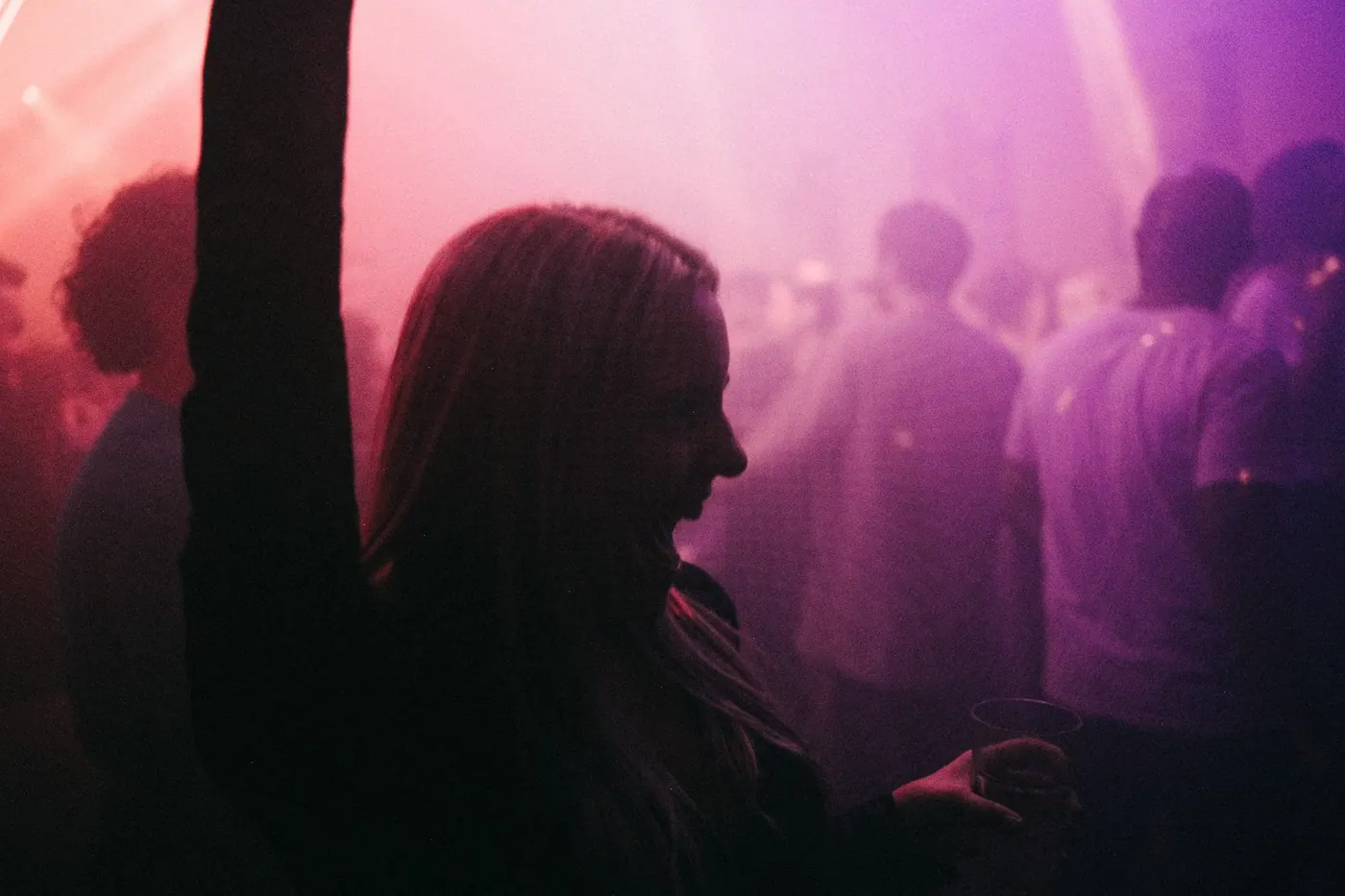 A woman dancing with her arm raised, silhouetted against pink and purple lights and a softly blurred crowd